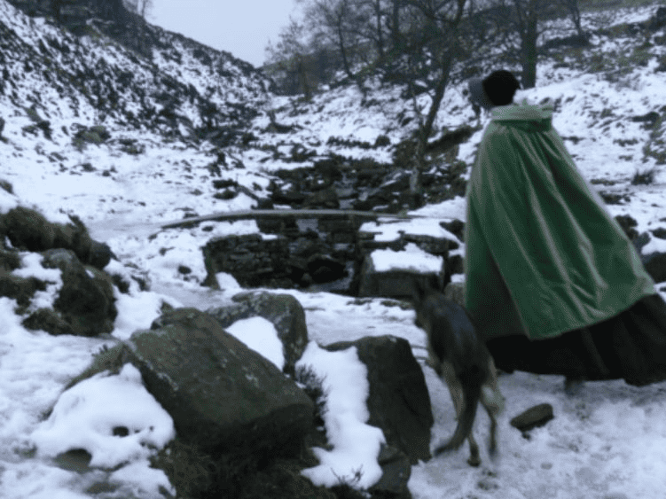 A woman wearing a green cape and looking like Jane Eyre would have is taking a walk in winter.