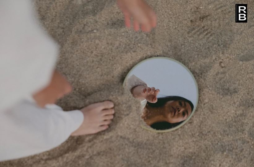A woman with her feet in the sand, seeing her reflection in a mirror.