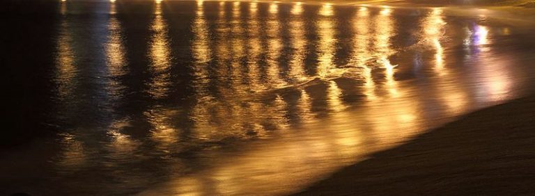 Beach at night, with lights reflected in the water.